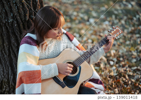 Young girl playing the guitar near the tree in autumn park. Beautiful evening shot of a girl musician outdoors in autumn nature Young girl playing the guitar near the tree in autumn park. Beautiful evening shot of a girl musician outdoors in autumn nature 139011418
