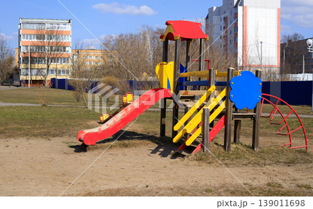 Wooden children's playground equipment with a red slide and blue climbing elements. Colorful playset in a residential area with apartment buildings Wooden children's playground equipment with a red slide and blue climbing elements. Colorful playset in a residential area with apartment buildings 139011698
