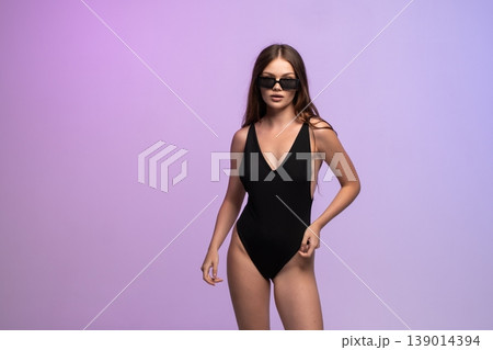 Confident young woman posing in black swimsuit and stylish sunglasses on purple gradient background Confident young woman posing in black swimsuit and stylish sunglasses on purple gradient background 139014394