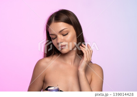 Young woman with closed eyes applying moisturizing facial cream, highlighting beauty, wellness, and a glowing complexion 139014428
