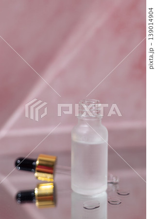 Cosmetic serum dropper with clear liquid and frosted glass bottle on pink reflective surface, skincare essence texture, beauty product concept Cosmetic serum dropper with clear liquid and frosted glass bottle on pink reflective surface, skincare essence texture, beauty product concept 139014904