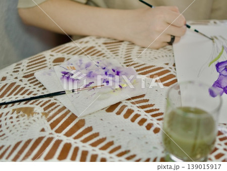 Artist's hands painting purple crocuses with watercolors. Creative workspace with palette and brushes, art education concept. Artist's hands painting purple crocuses with watercolors. Creative workspace with palette and brushes, art education concept. 139015917