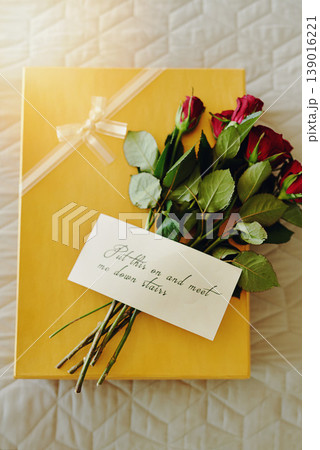 Box, roses and letter on bed, gift and invitation in closeup, above or message for valentines day in hotel. Flowers, bouquet or plants with cardboard package, notes or clothes in bedroom in home 139016221