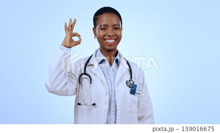 Portrait, okay or happy black woman doctor with smile or confidence isolated on blue background. Wellness, studio or female African healthcare worker with approval sign, success or yes hand gesture Portrait, okay or happy black woman doctor with smile or confidence isolated on blue background. Wellness, studio or female African healthcare worker with approval sign, success or yes hand gesture 139016675