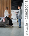 Two businesswomen greeting and talking in front of gray sofa in open modern office. Greeting, discussion, office, teamwork, business. 139020048