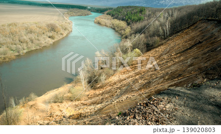 A river runs under a steep bank in a natural landscape. Trees line the river, and a forest is visible in the background. The scene is bright and shows a clear sky. 139020803