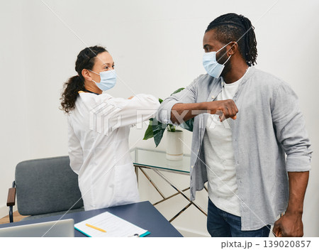 African American man and his doctor wearing medical masks greeting each other by elbow bumping at doctor's office. Elbow bump, battle coronavirus epidemic African American man and his doctor wearing medical masks greeting each other by elbow bumping at doctor's office. Elbow bump, battle coronavirus epidemic 139020857