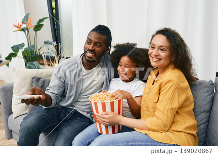Fun family with little daughter are watching comedy movie together and eating popcorn from a large striped bucket at cozy home 139020867