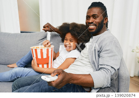 African American father with his little daughter hugging and watching cinema together and eating popcorn from a large striped bucket. Father's day African American father with his little daughter hugging and watching cinema together and eating popcorn from a large striped bucket. Father's day 139020874