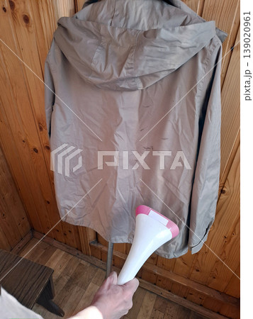 A person uses a handheld garment steamer to smooth out wrinkles on a beige hooded jacket. The jacket hangs against a wooden wall in a cozy indoor space. A person uses a handheld garment steamer to smooth out wrinkles on a beige hooded jacket. The jacket hangs against a wooden wall in a cozy indoor space. 139020961