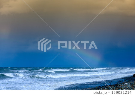 A vibrant rainbow arches over stormy waves at the beach during a dramatic sunset sky A vibrant rainbow arches over stormy waves at the beach during a dramatic sunset sky 139021214