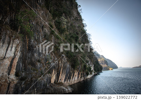 Dramatic rock cliff along calm mountain lake under clear sky, scenic wilderness landscape with textured stone wall and copy space. 139022772