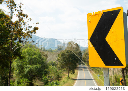 Yellow left curve warning road sign on rural asphalt road, mountain landscape and trees under bright daylight travel scene. 139022938