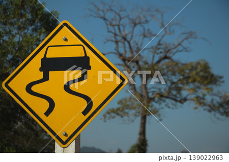 Yellow slippery road warning sign with skidding car symbol on rural roadside under clear blue sky. Yellow slippery road warning sign with skidding car symbol on rural roadside under clear blue sky. 139022963