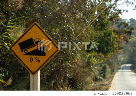 Steep 8 percent grade warning sign along a narrow rural mountain road surrounded by dense green vegetation in daylight. Steep 8 percent grade warning sign along a narrow rural mountain road surrounded by dense green vegetation in daylight. 139022968