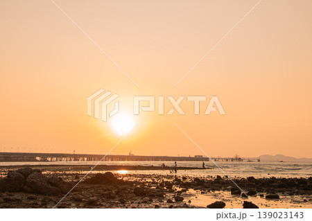 Warm sunlight fades over a rocky beach as distant figures explore the shoreline near a long pier, capturing a tranquil seaside moment with glowing reflections and a soft horizon. Warm sunlight fades over a rocky beach as distant figures explore the shoreline near a long pier, capturing a tranquil seaside moment with glowing reflections and a soft horizon. 139023143