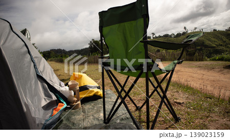 Camping setup with folding chair, tent and outdoor gear on grassy ground, countryside landscape under cloudy sky, peaceful travel and adventure lifestyle concept. Camping setup with folding chair, tent and outdoor gear on grassy ground, countryside landscape under cloudy sky, peaceful travel and adventure lifestyle concept. 139023159
