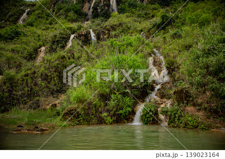 Small natural waterfall flowing through lush green hillside into a clear pool. Tropical forest landscape with fresh water stream and tranquil outdoor environment. 139023164