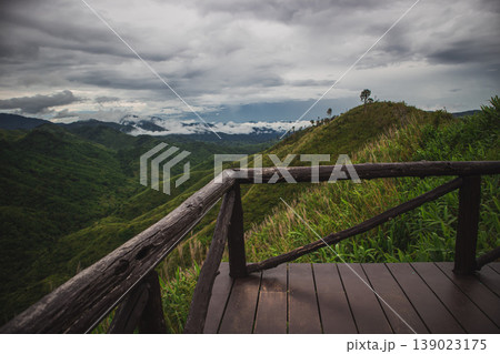 Wooden viewpoint deck overlooking lush green mountain ridge and misty valley under dramatic cloudy sky. Scenic highland panorama, travel destination and nature adventure concept. Wooden viewpoint deck overlooking lush green mountain ridge and misty valley under dramatic cloudy sky. Scenic highland panorama, travel destination and nature adventure concept. 139023175