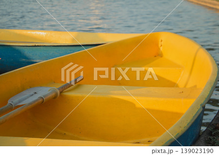 Yellow rowboat with oar resting by calm water under warm sunlight. Minimal outdoor scene with copy space, ideal for travel, summer leisure, relaxation and waterfront themes. 139023190