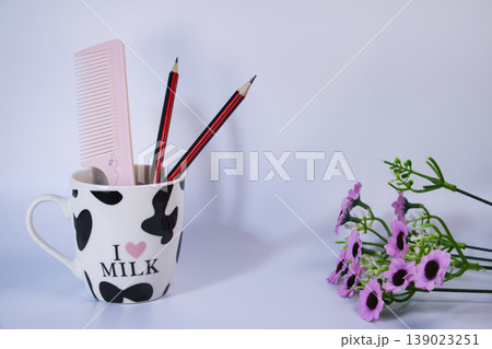 Pencils and pink comb in ceramic cup with small purple flowers on white background. Minimal still life workspace or study concept with copy space. Pencils and pink comb in ceramic cup with small purple flowers on white background. Minimal still life workspace or study concept with copy space. 139023251