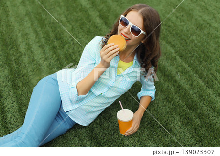 Happy young woman eating burger fast food drinking juice, teenage girl lying in summer park Happy young woman eating burger fast food drinking juice, teenage girl lying in summer park 139023307