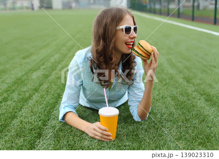 Happy young woman eating burger fast food drinking juice, teenage girl lying in summer park 139023310