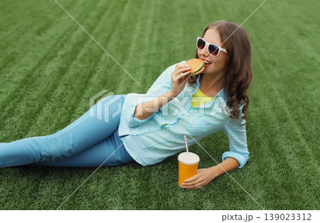 Happy young woman eating burger fast food drinking juice, teenage girl lying in summer park 139023312