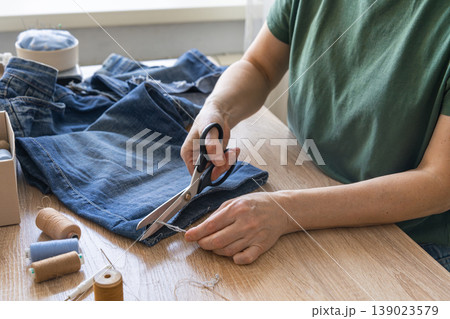 Hands cutting thread from old jeans for reuse, promoting sustainable fashion and circular economy principles. Person upcycling denim jeans with scissors, repairing clothing 139023579
