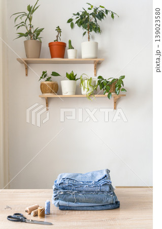 Stack of old blue jeans ready for upcycling on a wooden table with scissors and thread. Potted plants decorating shelves in background. Reusing things. Environmental awareness Stack of old blue jeans ready for upcycling on a wooden table with scissors and thread. Potted plants decorating shelves in background. Reusing things. Environmental awareness 139023580