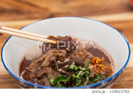 Thai boat noodle soup with beef slices meatball herbs and dark broth held by chopsticks in bowl on wooden table showing rich flavor and traditional street food style Thai boat noodle soup with beef slices meatball herbs and dark broth held by chopsticks in bowl on wooden table showing rich flavor and traditional street food style 139023782