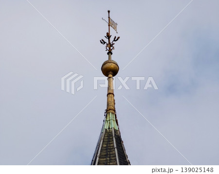 Slim Spire With Gleaming Ornament. Refined Vertical Spire Featuring Gilded Cross Against Cloudy Sky 139025148