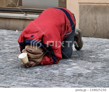 Beggar On Cold City Street. Depiction Of Downcast Individual Asking For Charity Amidst City Streets Beggar On Cold City Street. Depiction Of Downcast Individual Asking For Charity Amidst City Streets 139025150