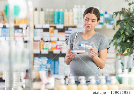 Attentive young pregnant girl buyer choosing medical ointment in pharmacy Attentive young pregnant girl buyer choosing medical ointment in pharmacy 139025988