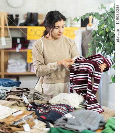 Latina female buyer standing in clothing market and carefully examines selection of sweaters on sale of winter clothing collection 139026033