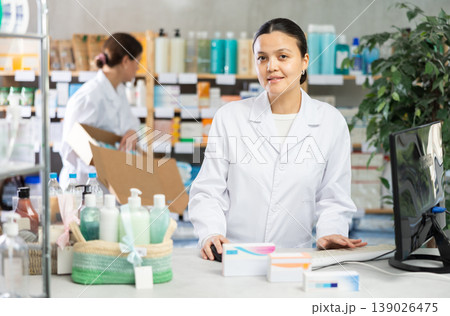 Portrait of young asian woman pharmacist standing at prescription counter and working on computer, stocktaking medicines at pharmacy shop and female assistant in background 139026475