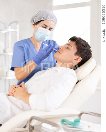 Closeup face of young male client receiving injections during lip enhancement procedure, professional cosmetologist hands in gloves holding syringe Closeup face of young male client receiving injections during lip enhancement procedure, professional cosmetologist hands in gloves holding syringe 139026516