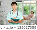 Smiling man beautician in uniform standing in cosmetology clinic, filling out clipboard with medical records 139027359