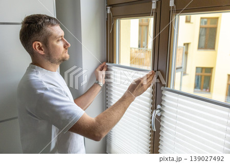 Man adjusting window blinds in modern apartment interior with soft natural light Man adjusting window blinds in modern apartment interior with soft natural light 139027492