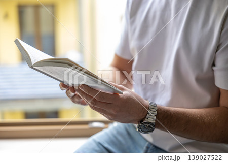 Man reading book sitting on window sill in bright minimal interior with natural light 139027522