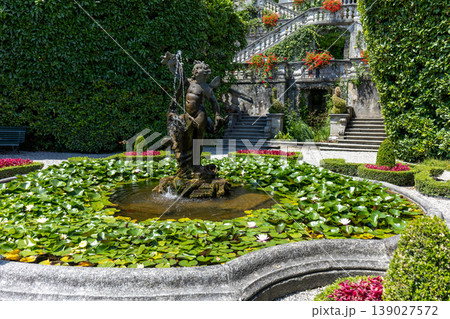 Decorative garden fountain with sculpture surrounded by flowers and classical architecture 139027572