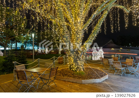 Outdoor festive lighting with illuminated tree and cozy seating area night holiday atmosphere Outdoor festive lighting with illuminated tree and cozy seating area night holiday atmosphere 139027626