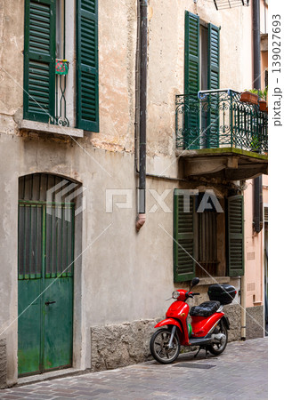A bright red scooter parked along a narrow European street with aged plaster walls and green shutters. 139027693
