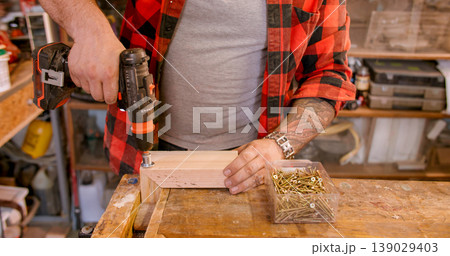Screw driving. Furniture assembly. Male carpenter hand with cordless electric drill connecting corner plank set on wooden workbench. Screw driving. Furniture assembly. Male carpenter hand with cordless electric drill connecting corner plank set on wooden workbench. 139029403