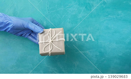 Gift delivery. Pandemic holidays. Hand in protective glove holding wrapped present box on blue textured background with copy space. 139029683