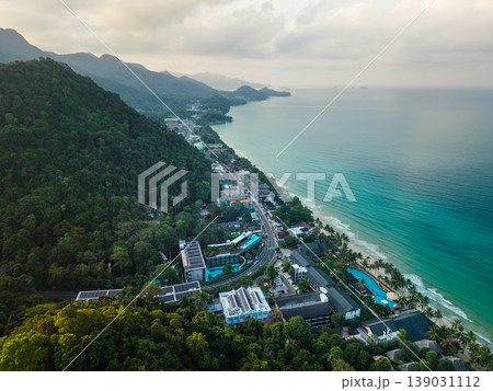 Aerial view of beachfront resorts, solar panels, and a coastal road winding through a lush tropical mountain island. Aerial view of beachfront resorts, solar panels, and a coastal road winding through a lush tropical mountain island. 139031112