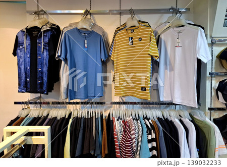 Yogyakarta, Indonesia - March 18th 2026: Rows of colorful T-shirts, a blue denim jacket, and various casual garments neatly arranged on metal hangers in a modern clothing store. Yogyakarta, Indonesia - March 18th 2026: Rows of colorful T-shirts, a blue denim jacket, and various casual garments neatly arranged on metal hangers in a modern clothing store. 139032233