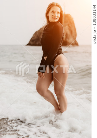 Swimsuit beach model posing in black swimwear among ocean waves and coastal rock formation during sunset golden hour Swimsuit beach model posing in black swimwear among ocean waves and coastal rock formation during sunset golden hour 139033144