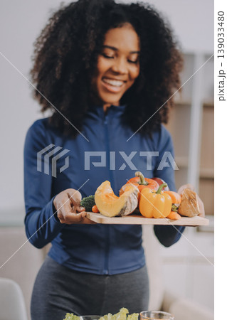 Young nutritionist showing bell peppers during an online video call consulting about healthy eating, sitting at home at a table with vegetables, laptop and measuring tape 139033480