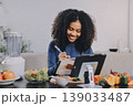 Young nutritionist showing bell peppers during an online video call consulting about healthy eating, sitting at home at a table with vegetables, laptop and measuring tape 139033487